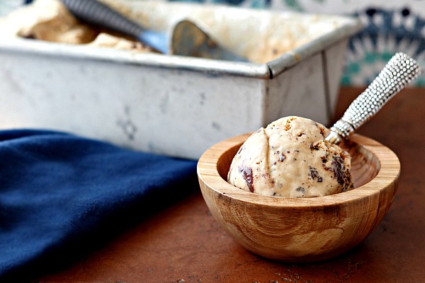 A metal loaf pan of salted caramel turtle fudge swirl  with an ice cream scoop and a wooden bowl with a serving of the ice cream.