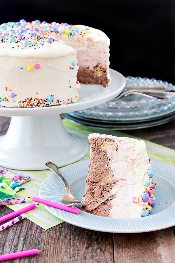 A chocolate, vanilla and strawberry ice cream birthday cake on a pedestal with a slice on a plate.