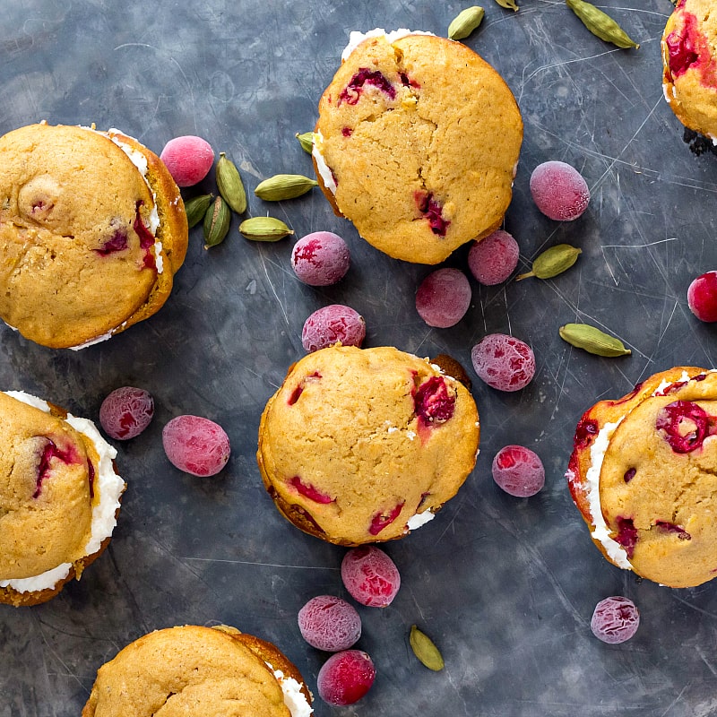 Overhead shot of filled pumpkin cranberry whoopie pies on a slate background with cardamom pods and fresh cranberries.