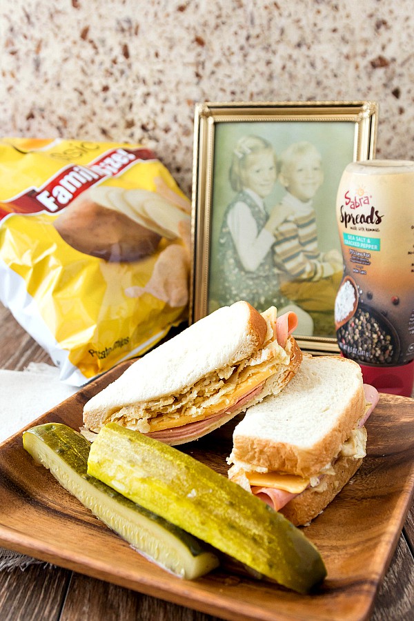 A bologna and cheese sandwich on a wooden plate with pickle spears and a bag of potato chips and a family photo in the background.