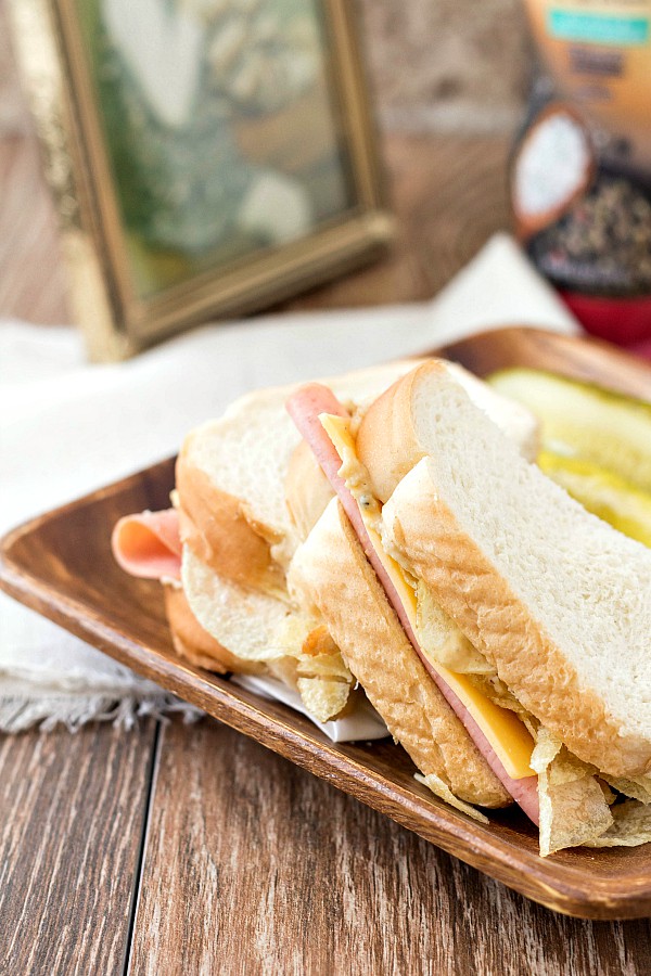 A bologna and cheese sandwich cut in half on a wooden plate with pickle spears.