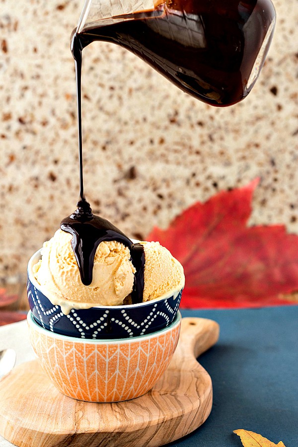 Orange Cardamom Pumpkin Ice Cream in a bowl with a clear glass pitcher of maple chocolate sauce pouring over it.