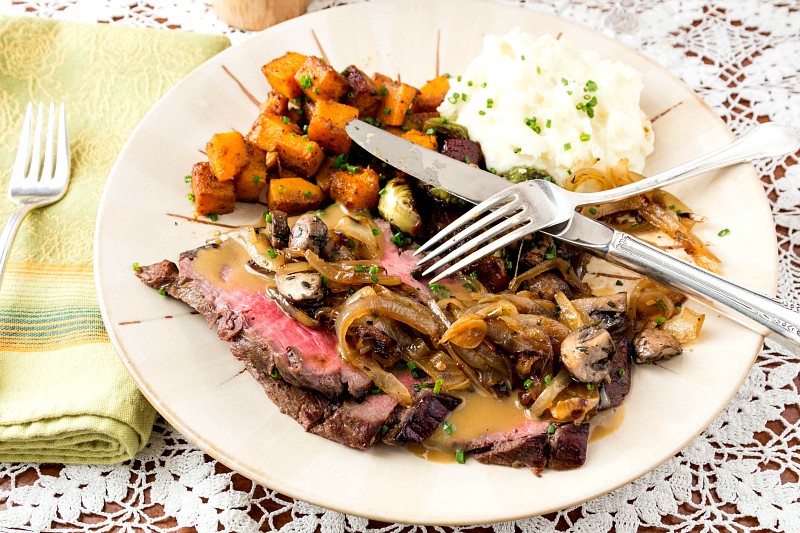 Sweet and Spicy Marinated London Broil with sauce on a white plate with caramelized onions and mushrooms, mashed potatoes, roasted butternut squash cubes and brussels sprouts with fork and knife crossed over plate.