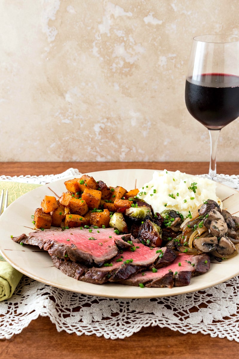 Sweet and Spicy Marinated London Broil from Pastry Chef Online served on a white plate with sauteed mushrooms, mashed potatoes, roasted butternut squash cubes and brussels sprouts with a glass of red wine.