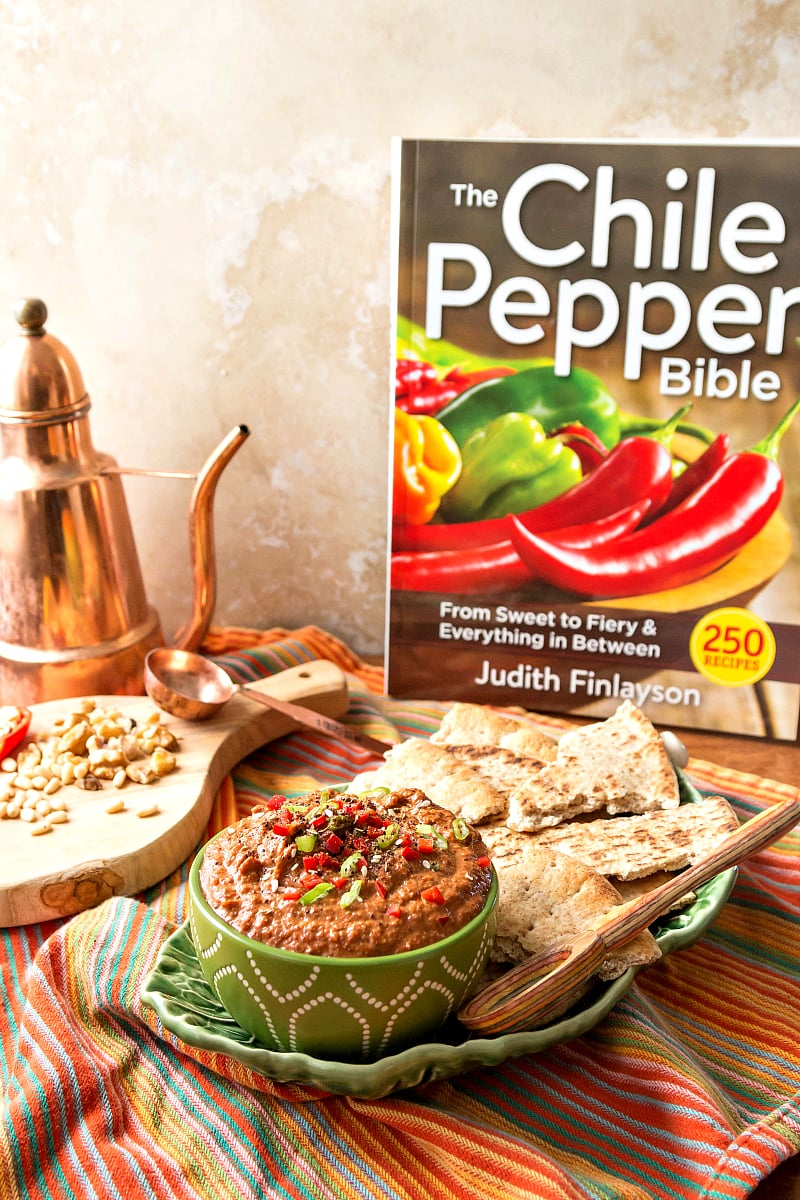 A green bowl of muhammara with pita, ready for serving and a cookbook, "The Chile Pepper Bible".