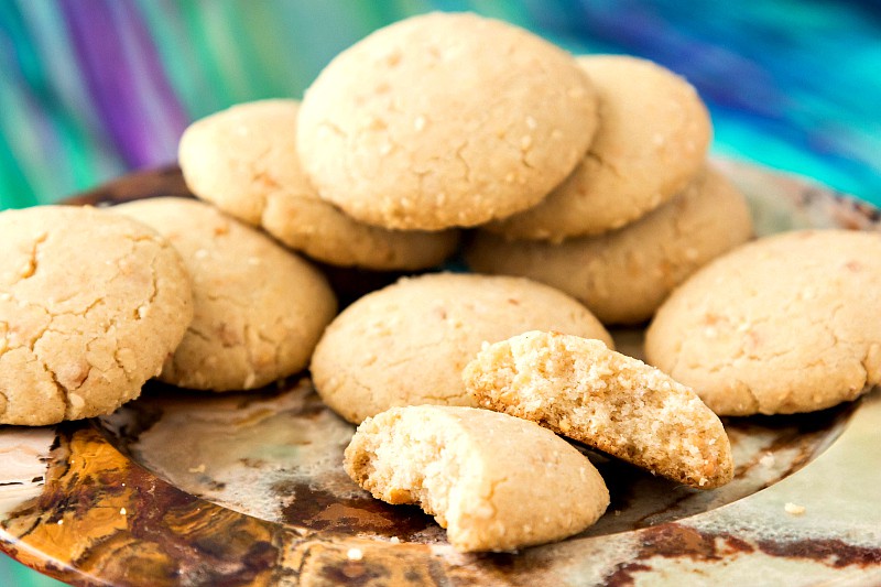 A pile of  ghoriba bahla cookies on a plate. One is broken in half to ese the crumbly shortbread like texture. The tops are light brown and cracked with almonds and sesame seeds showing in a few places. 