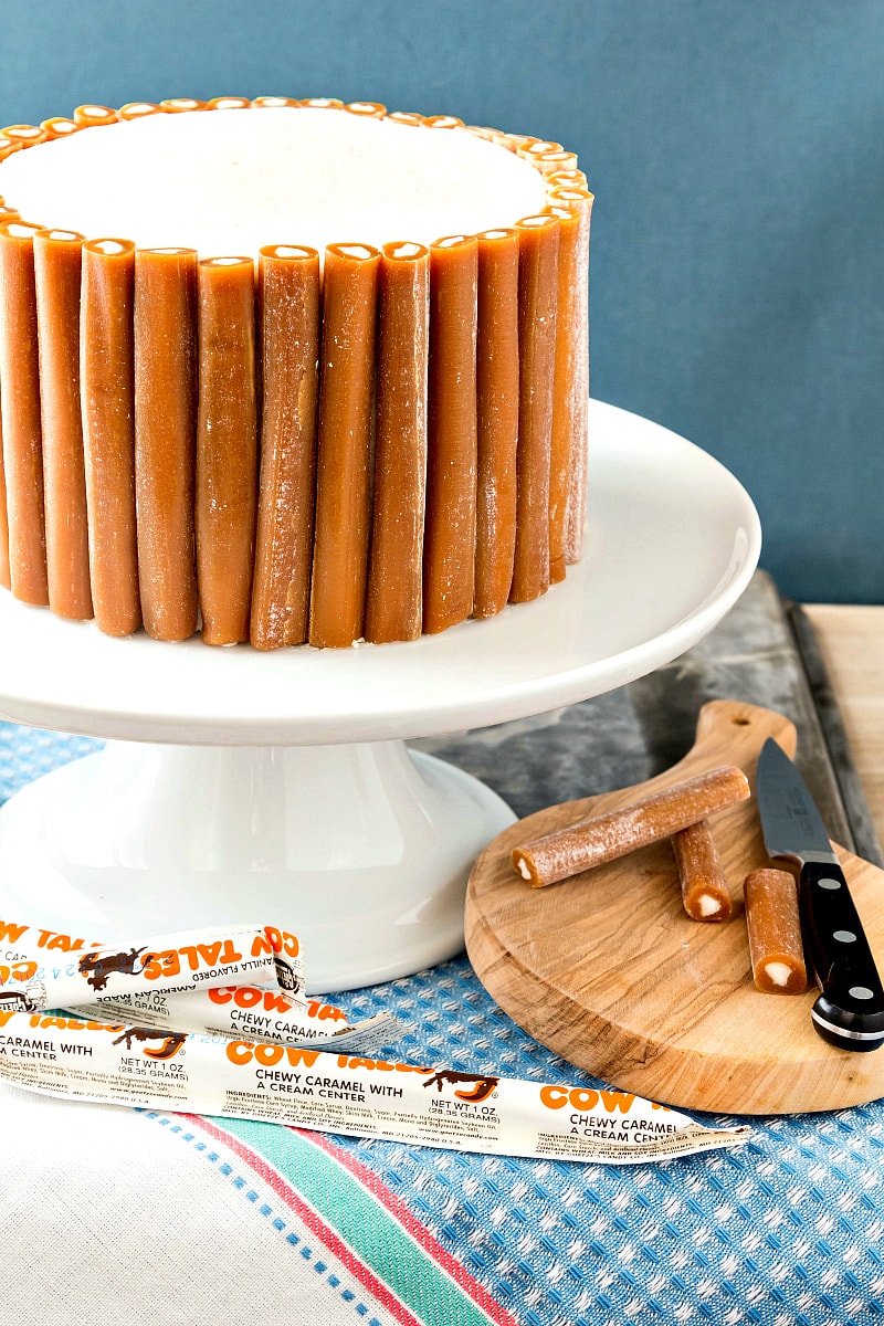 A cake decorated with caramel candies all around the sides on a white cake stand with Cow Tales candies in front.