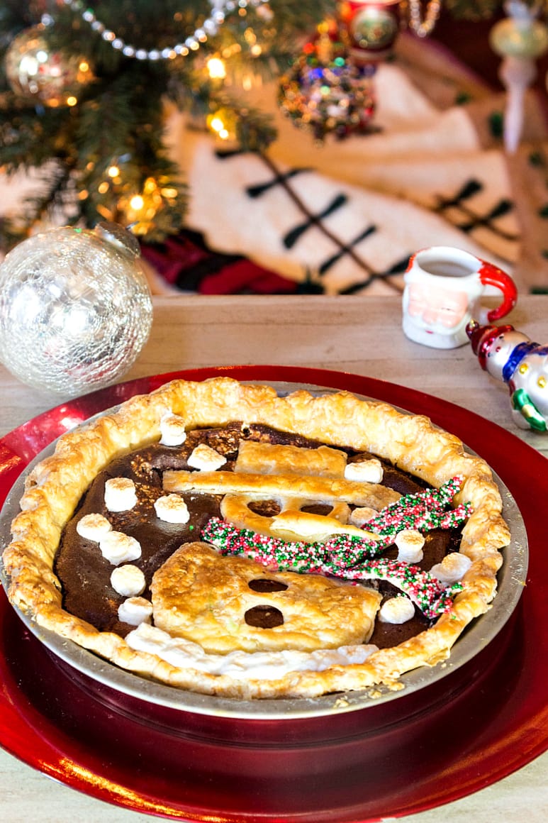 A whole pie with a snowman crust applique in a pie pan with a Christmas tree in the background.