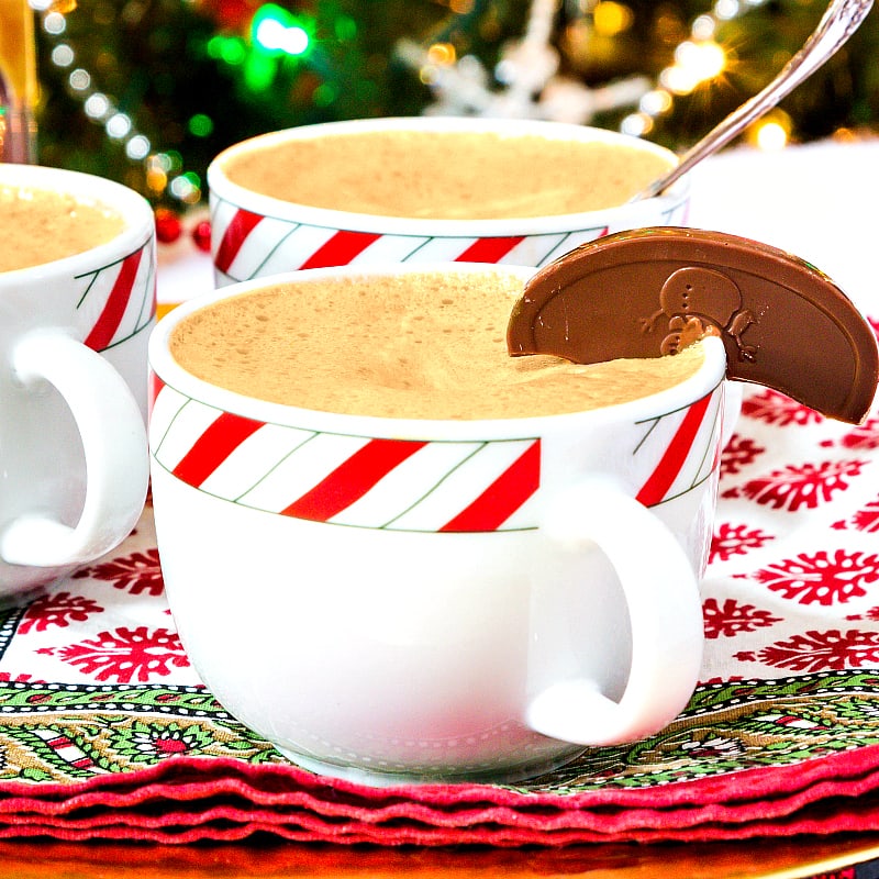 3 mugs of orange chocolate drinking chocolate. Each is a holiday themed mug with a band of red, white and green around the top. They are sitting on red and green napkins. There is a semi-cirle of chocolate garnishing the edge of the front mug. Christmas tree and lights are in the background.