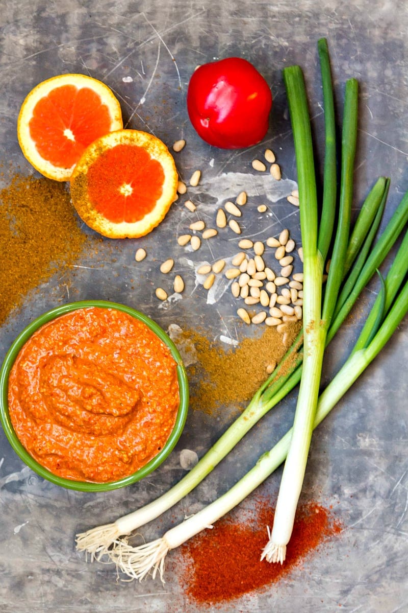 A bowl of blood orange muhammara, slices of blood orange, a red pepper, green onions, pine nuts and spices on a tray.