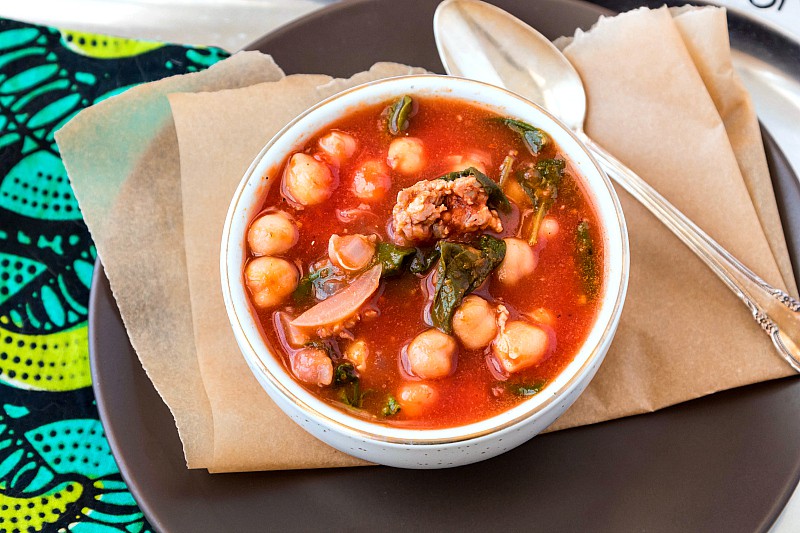 A bowl of tomato-based soup with Italian sausage, spinach and chickpeas with a silver spoon.