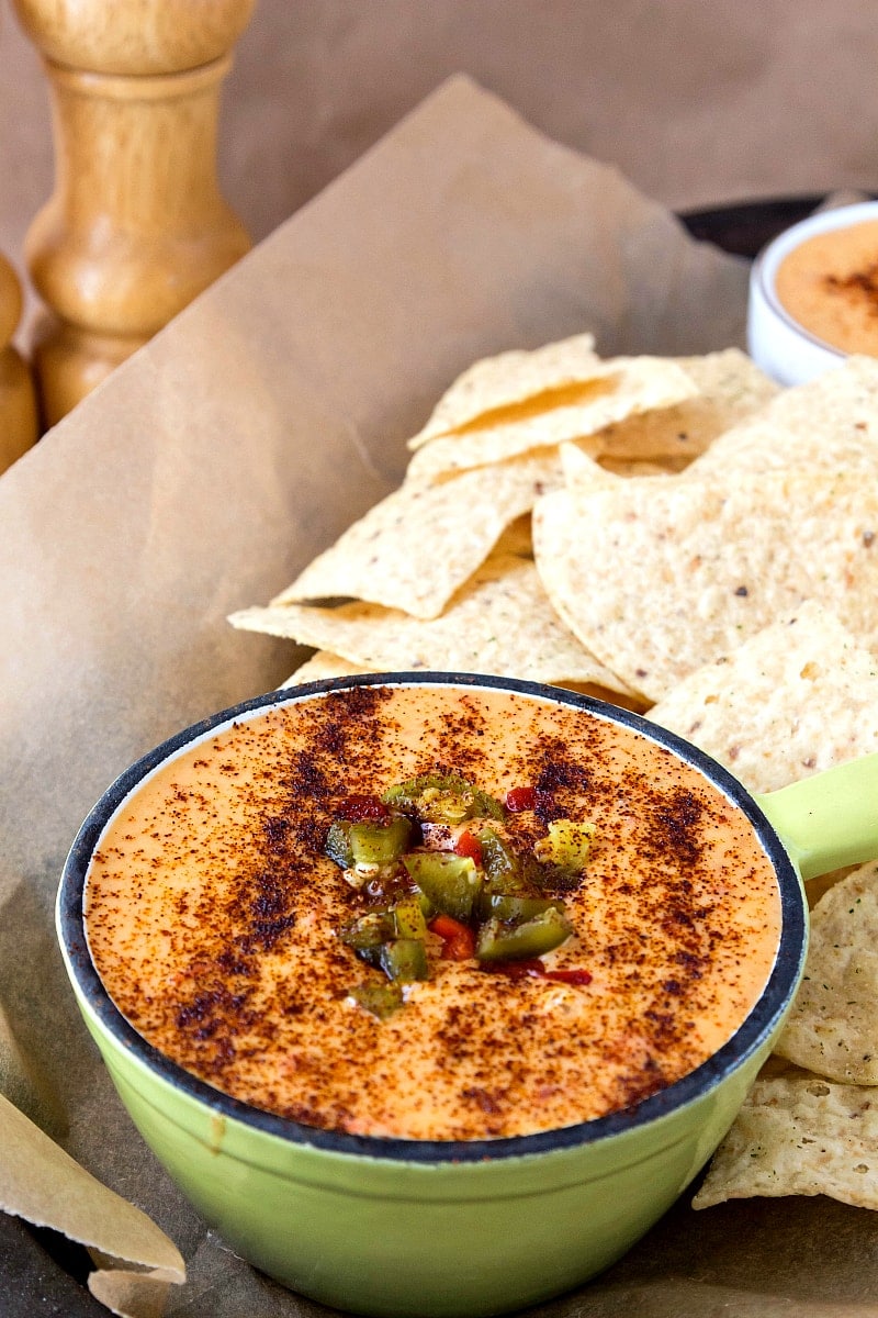Spicy cheese dip in a green ramekin topped with salsa, diced jalapenos and chipotle powder along with chips ready for serving.
