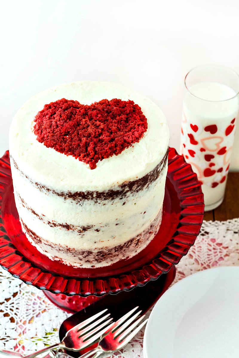 3/4 view of tall stacked red velvet cake frosted in white with a red heart on top