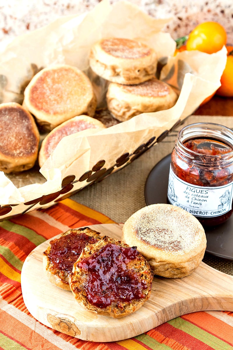 A split and toasted sprouted English muffin with butter and jam with a basket of English muffins and a jar of fig jam in the background.