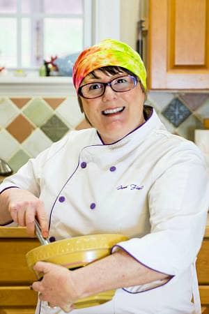 A woman in a tie-dyed bandana and a white chef coat smiling at the camera and holding a bowl with a whisk.