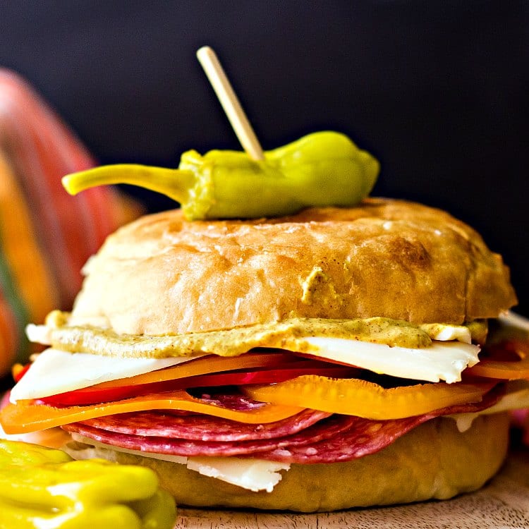 A sandwich made on a potato roll with cold cuts, sliced peppers, cheese, and mustard.