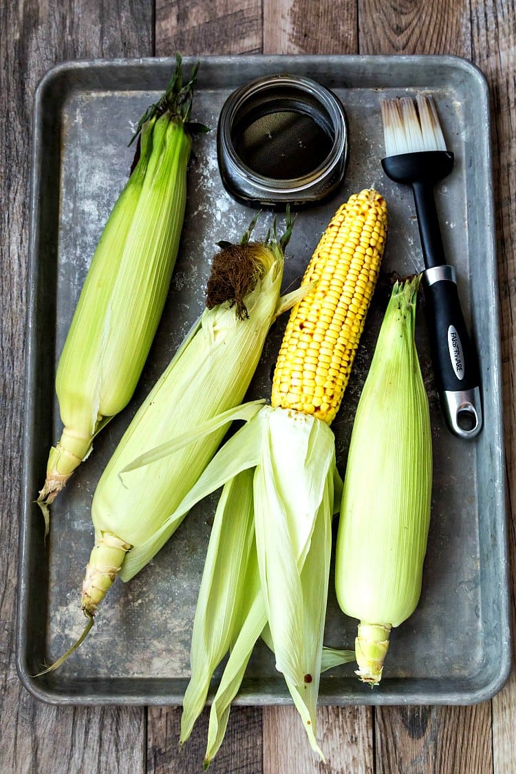 4 ears of corn with husks on a metal baking tray. One of the ears of corn has the husk pulled back so you can see the corn. Tea reduction and brush are ready to coat corn.