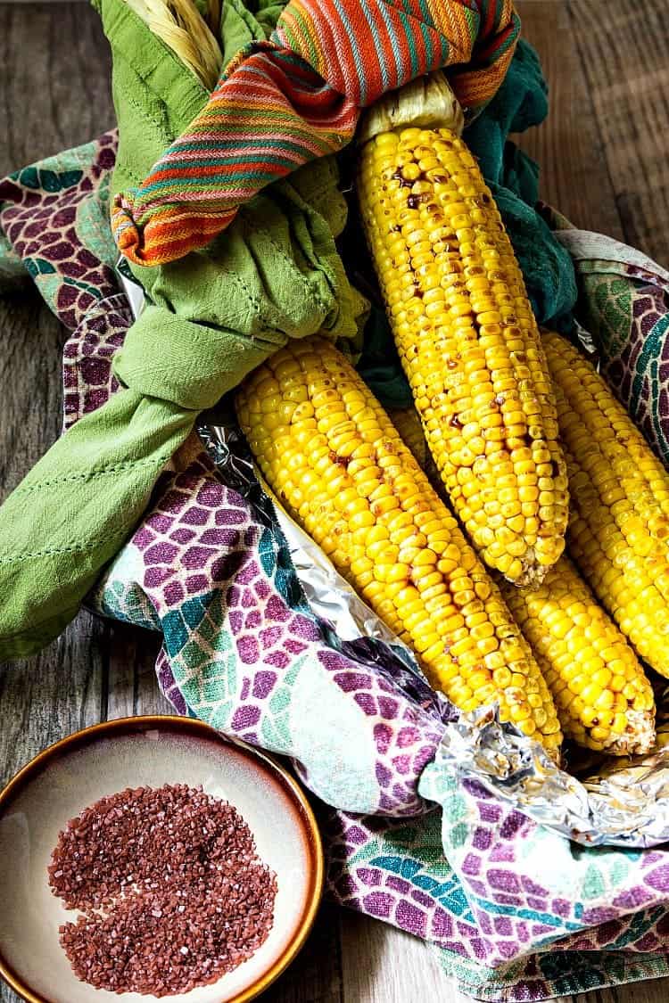 3 whole ears of roasted corn in a basket with a small plate of pink salt to serve with it.