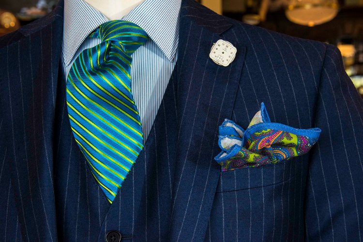 A blue pinstriped suit on a mannequin with blue, yellow and green striped neck tie, paisley, handkerchief and watch lapel pin.