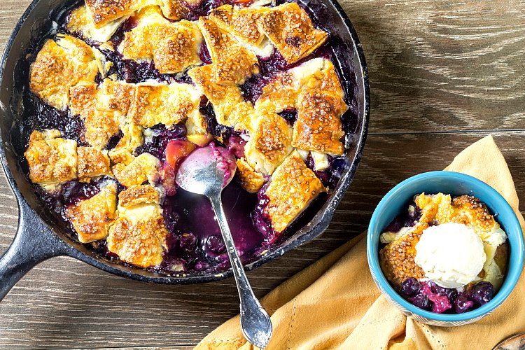 A horizontal shot of a cast iron skillet with cooked fruit and pieces of pie crust in it with a bowl of dessert scooped out with ice cream on top.