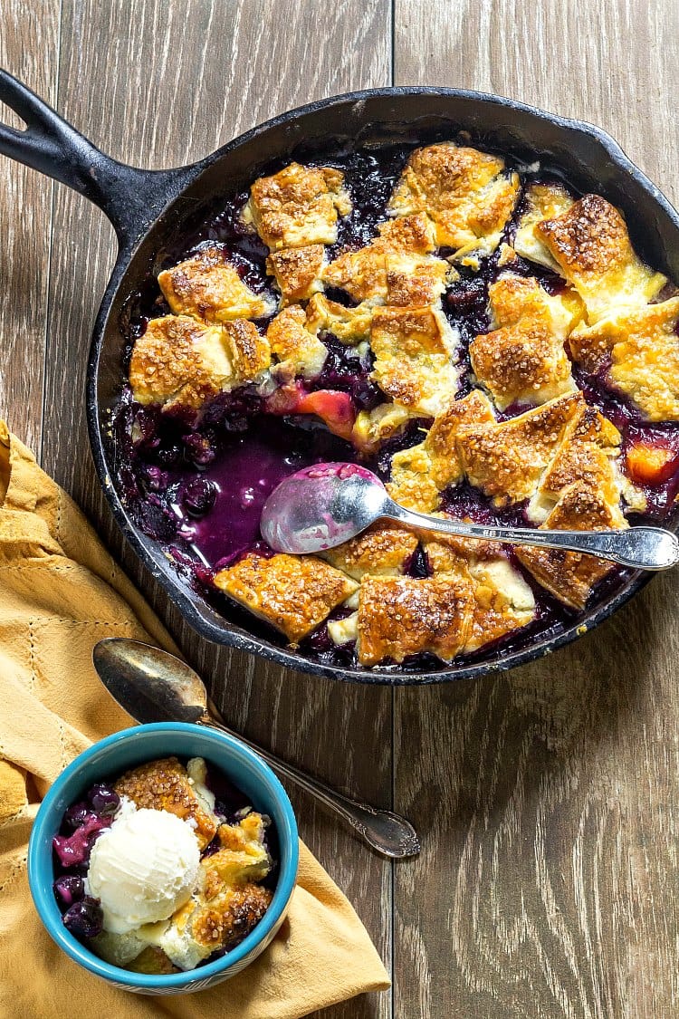 Overhead shot of pandowdy in a cast iron skillet with a bowl of blueberry peach pandowdy topped with vanilla ice cream.