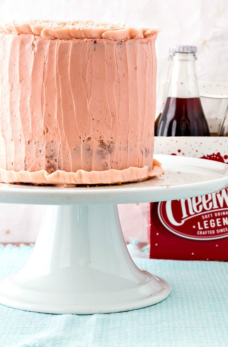 Side view of a pink-iced cake on a white cake stand with a 4 pack of cherry soda in the background.