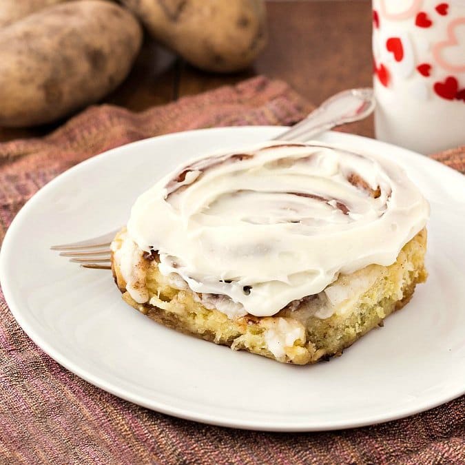 A whole iced gooey cinnamon roll on a white plate with a fork and a glass of milk on the side.