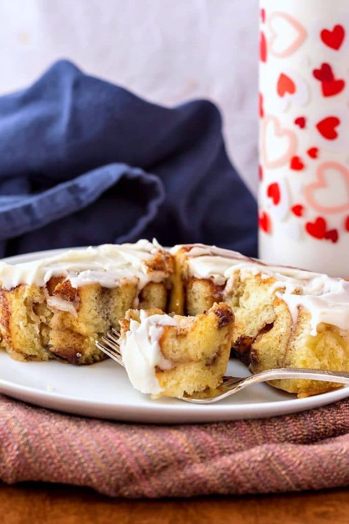 Cut and iced cinnamon roll on a white plate with a fork and a glass of milk in the background.