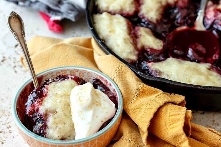 A bowl of cherry blueberry cobbler with hand whipped cream on top. Also a sprinkle of sugar in the raw on top.