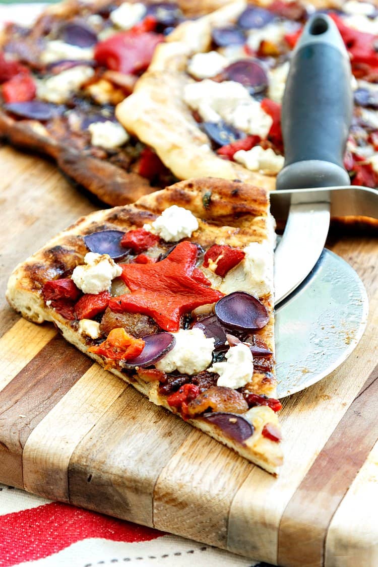 A slice of grilled pizza with goat cheese, sliced blue potatoes, and roasted red pepper. One of the peppers is cut in the shape of a star.