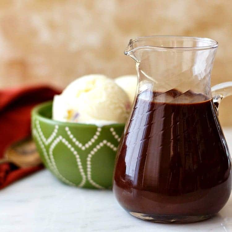 Square image of ice cream in a bowl and a pitcher of hot fudge ice cream sauce.