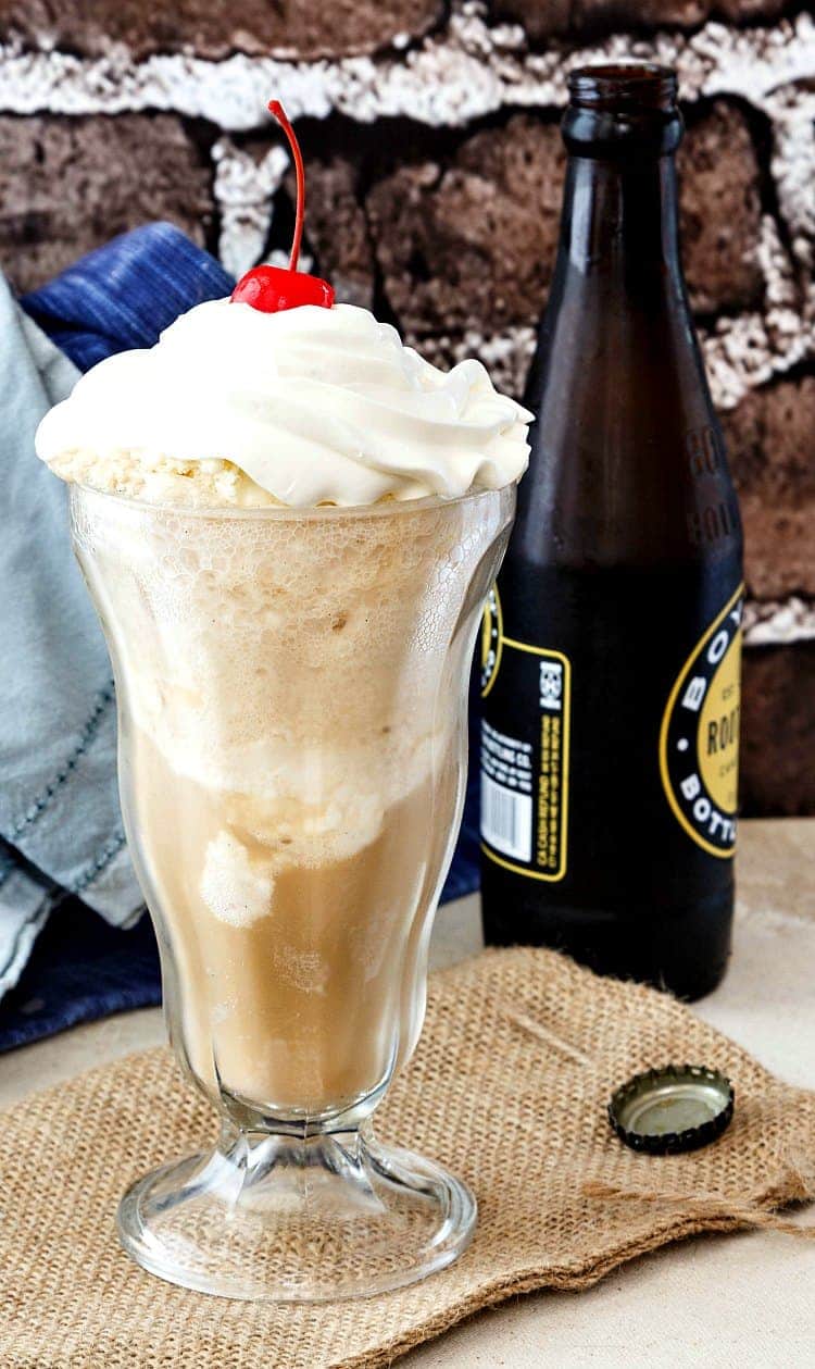 A tall glass of root beer float with a cherry on top and a bottle of root beer next to it.