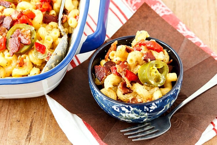 A closeup of a serving of southern mac and cheese topped with crumbled bacon, sweet red peppers, and pickled jalapeno in a small blue bowl with a fork ready for eating.