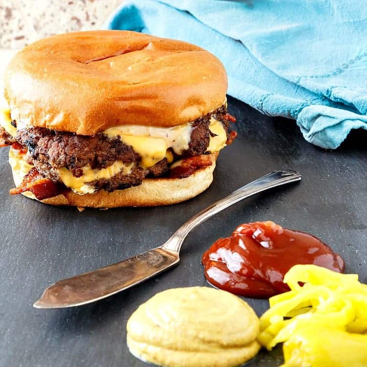 Square image of a bacon double cheeseburger with mustard, ketchup, and some pepperoncini.