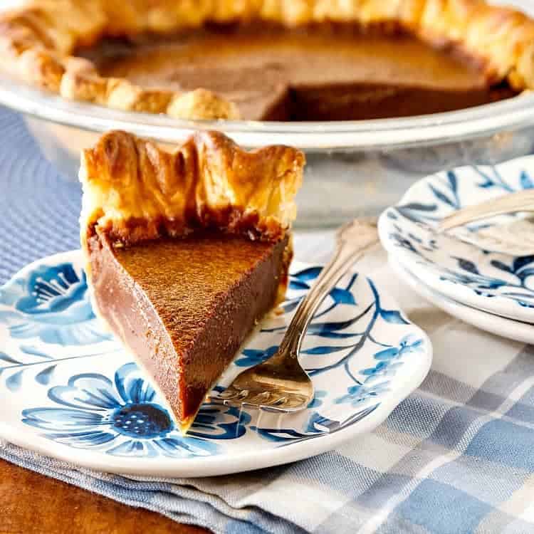 A slice of buttermilk pie on a blue print plate with a silver fork. The rest of the pie in the background.