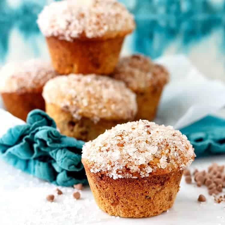 Muffins stacked on an aqua napkin with one in the front, ready for serving, wiht cinnamon chips scattered around.