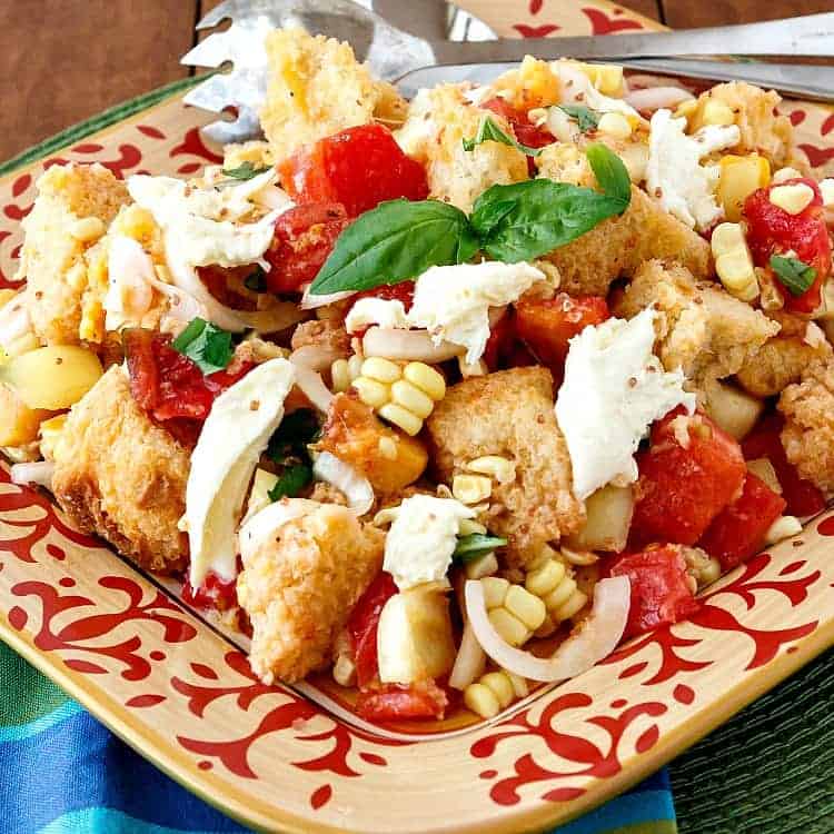 A square platter heaped with tomatoes, peaches, corn, basil, mozzarella cheese, and cubed bread.