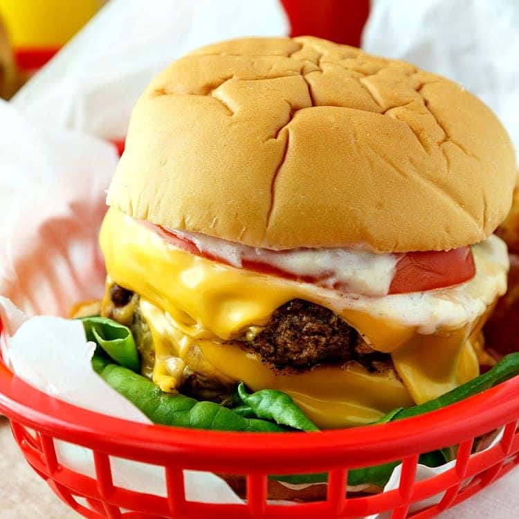 Close up of a  Double Polar Burger with Everything showing melting cheese, a slice of tomato, a hint of mayonnaise and fresh green lettuce. All sit on parchment paper in a red plastic basket.