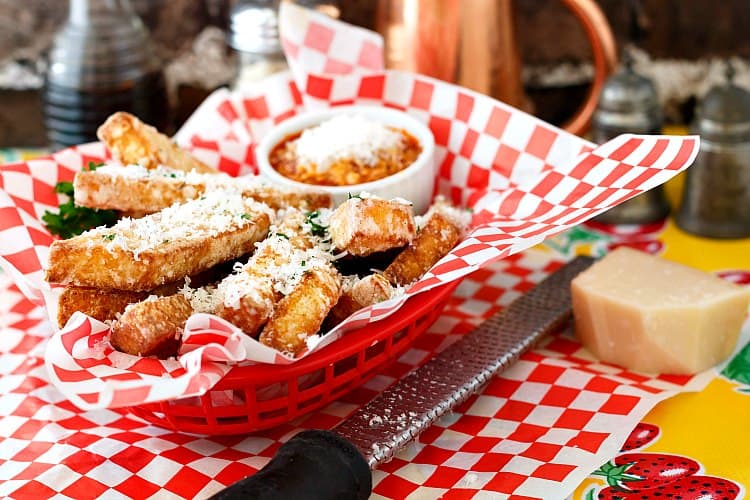 Full basket of fried polenta and dipping sauce all covered with grated Parmesan cheese. Microplane grater and a chunk of parmesan are to the side.