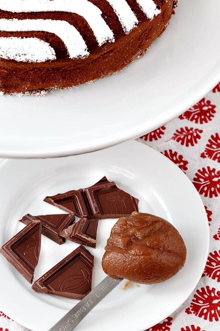A whole chestnut chocolate torte next to a plate with pieces of chocolate and a spoonful of chestnut puree.