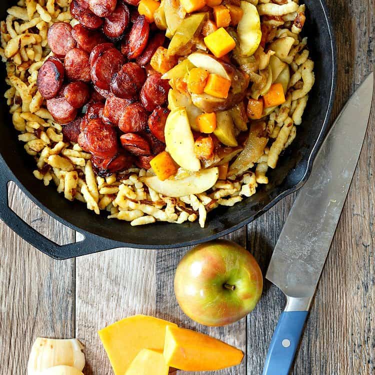 Crisped Polish sausage coins with sautéed apples, onions and butternut squash over spaetzle in a cast iron frying pan. Pan is on a wooden table. A large chef's knife is next to it as is a whole apple.  