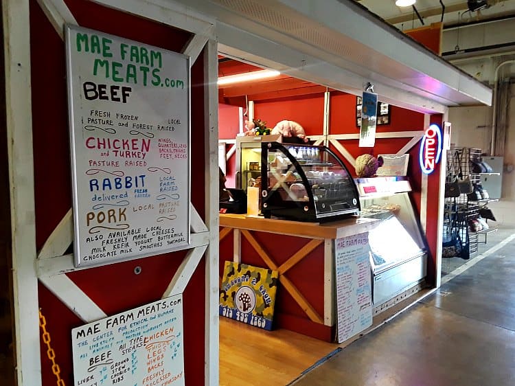 The MAE Farm stall at the state farmers market in Raleigh, NC. Red barn like wall, front glass counter, neon open sign. White board lists Beef, Chicken and Turkey, Rabbit and Pork.
