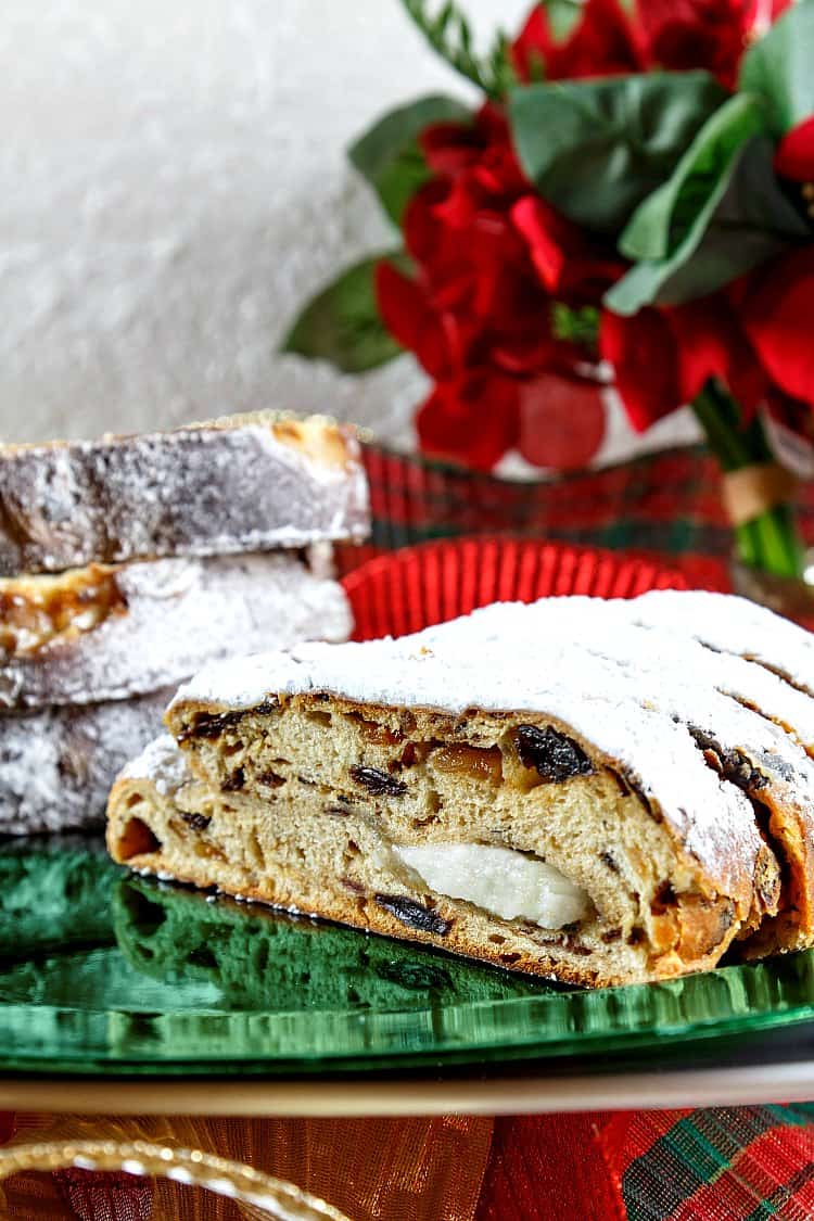 Cut open loaf of Christmas stollen showing dried fruits. Served on a bright green platter with a poinsettia in the background.