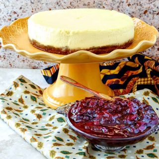 A glass dish of cranberry blueberry compote with a pale, creamy cheesecake on an orange cake stand in the background.