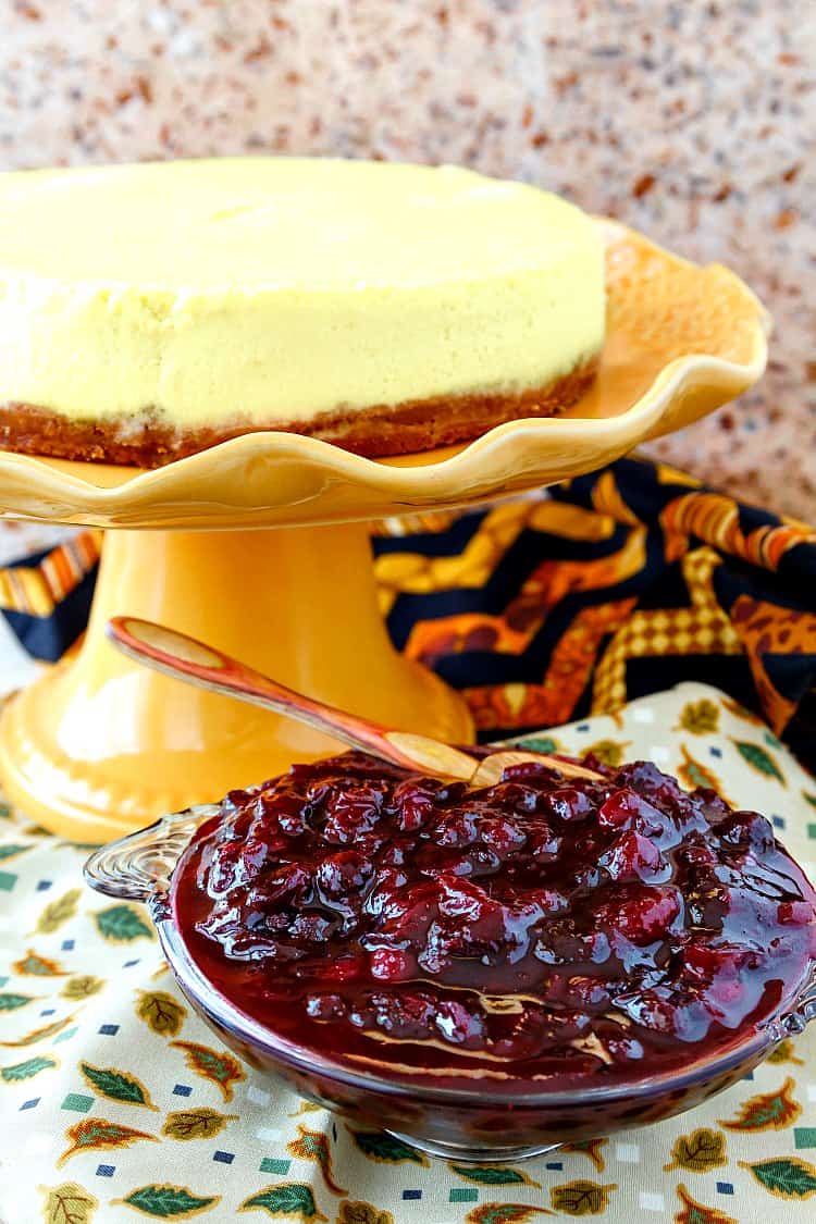 A whole roasted corn cheesecake on an orange footed cake stand with a bowl of cranberry blueberry compote next to it.
