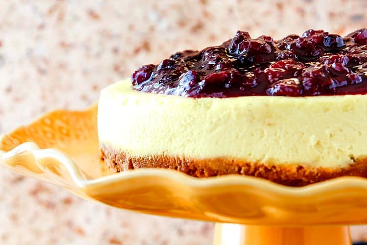 Close up view of the side of a cheesecake with fruit topping, sitting on an orange fluted cake stand.