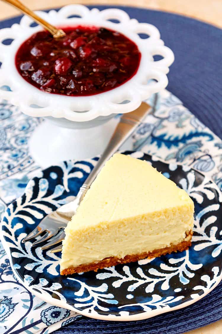 A slice of roasted corn cheesecake on a blue patterned plate with a white bowl of cranberry blueberry compote.