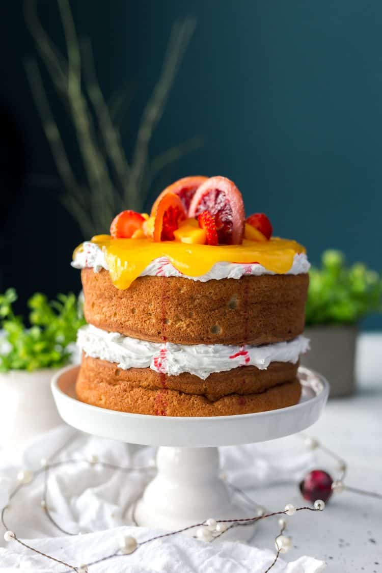 two layer sponge cake on white cake pedestal with whipped cream as filling and topping and decorated with orange slices and orange curd