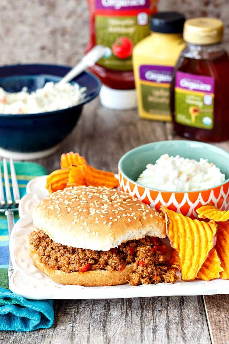 A sloppy joe on a sesame seed bun with coleslaw and ruffled chips as side dishes.