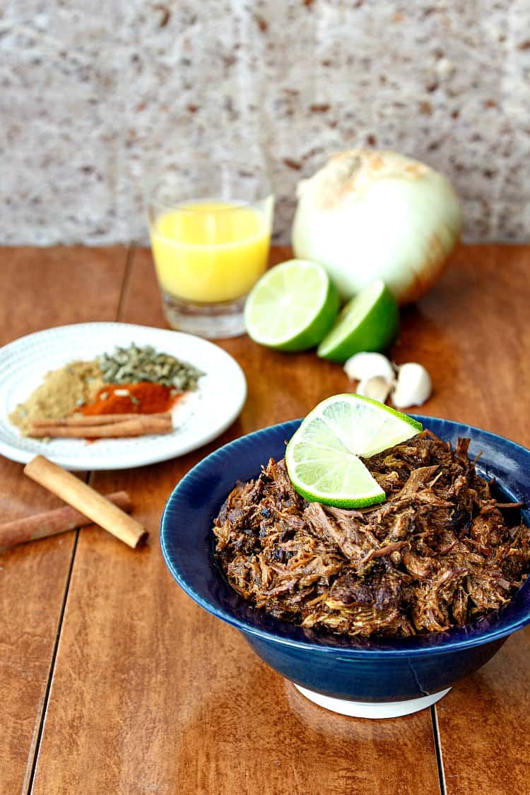 Shredded beef carnitas in a blue bowl, a plate of spices, and orange juice, cut lime, and an onion in the background.